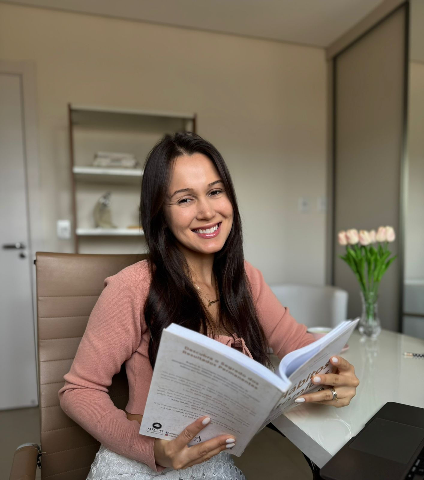 Andressa Guimarães sorrindo com um livro em seu ambiente de trabalho